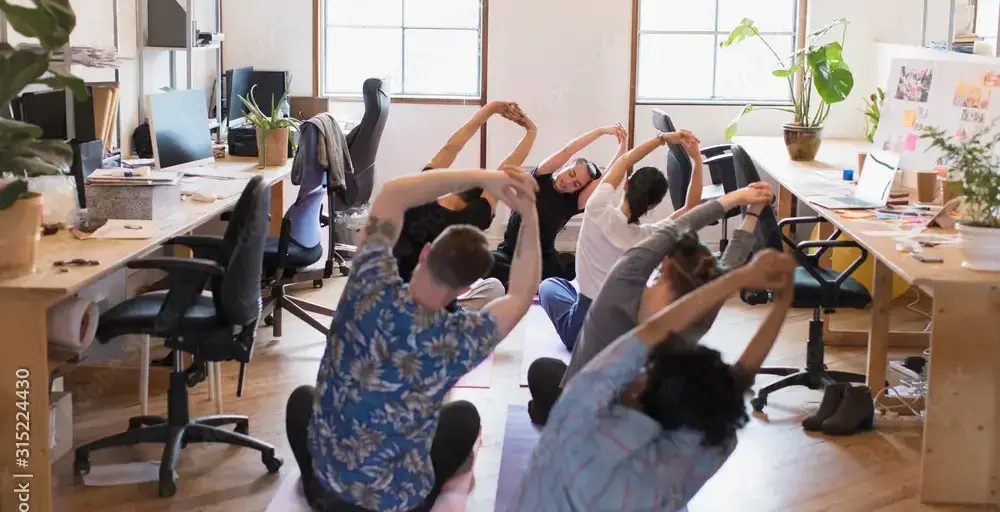 Group of coworkers on yoga mats doing a stretch in a bright open-plan office with desks and plants.