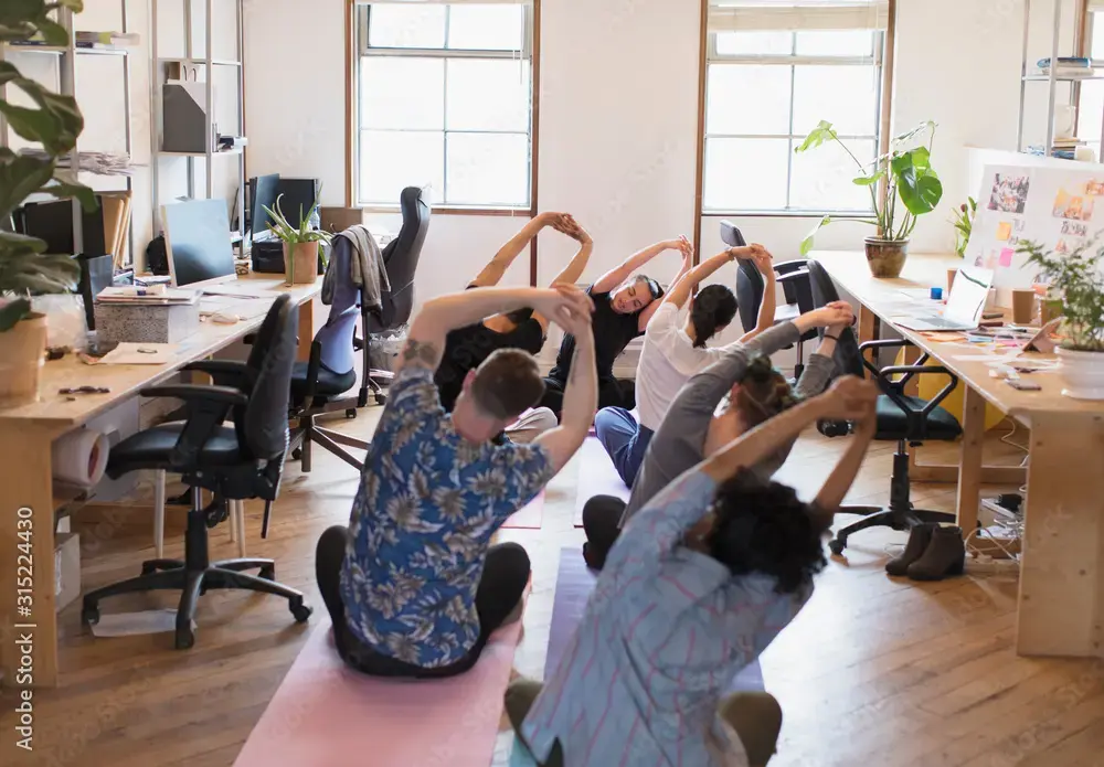 Group of coworkers on yoga mats doing a stretch in a bright open-plan office with desks and plants.