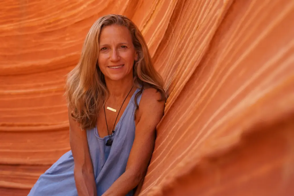 Smiling woman with long blond hair wearing a blue sleeveless top, standing against orange striped rock formations.