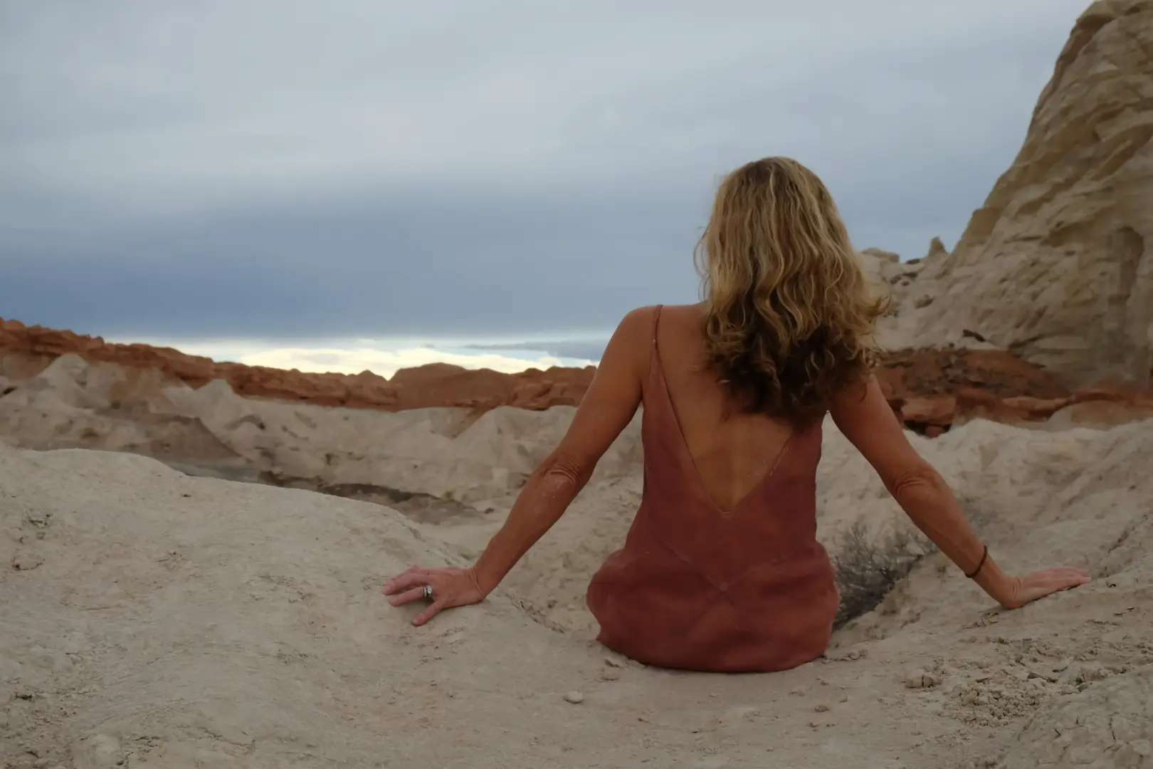 Person with long blond hair sits on light rock formations, seen from behind, wearing a reddish dress in a desert landscape.