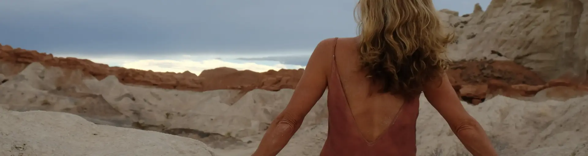 Person with long blond hair sits on light rock formations, seen from behind, wearing a reddish dress in a desert landscape.