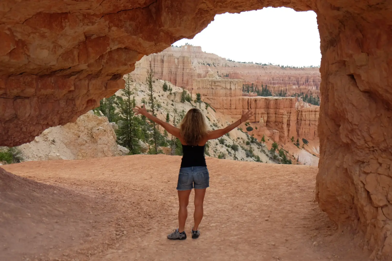 Person with arms outstretched stands on a dirt path inside a red canyon, surrounded by tall, eroded rock walls and distant cliffs.