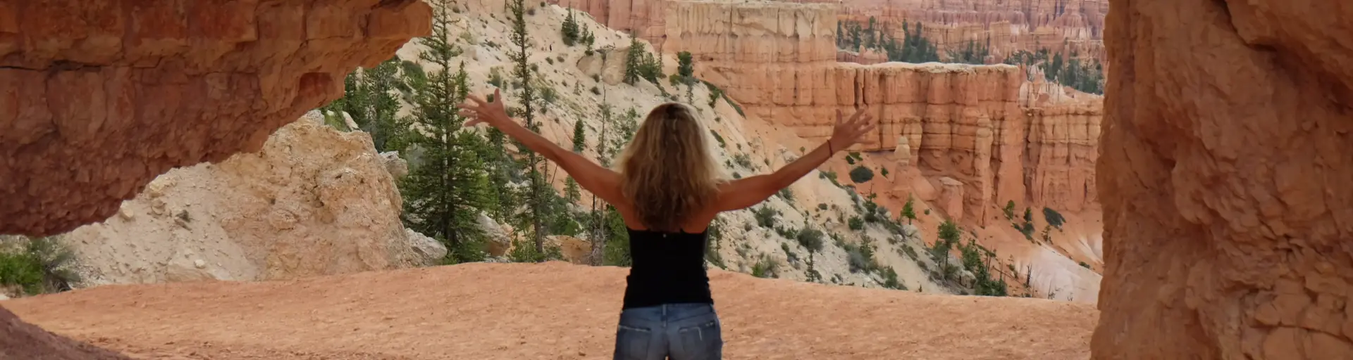 Person with arms outstretched stands on a dirt path inside a red canyon, surrounded by tall, eroded rock walls and distant cliffs.
