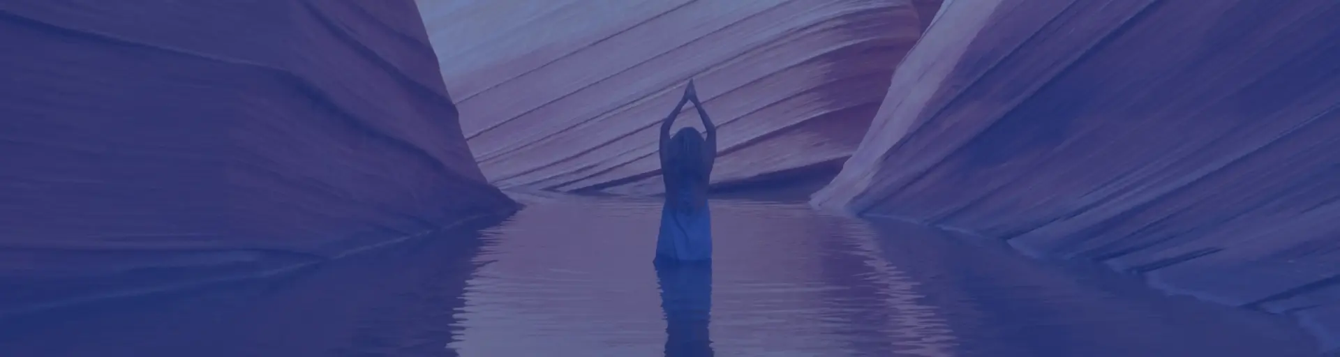 Person standing in shallow water between canyon walls with arms raised above head in a yoga pose.