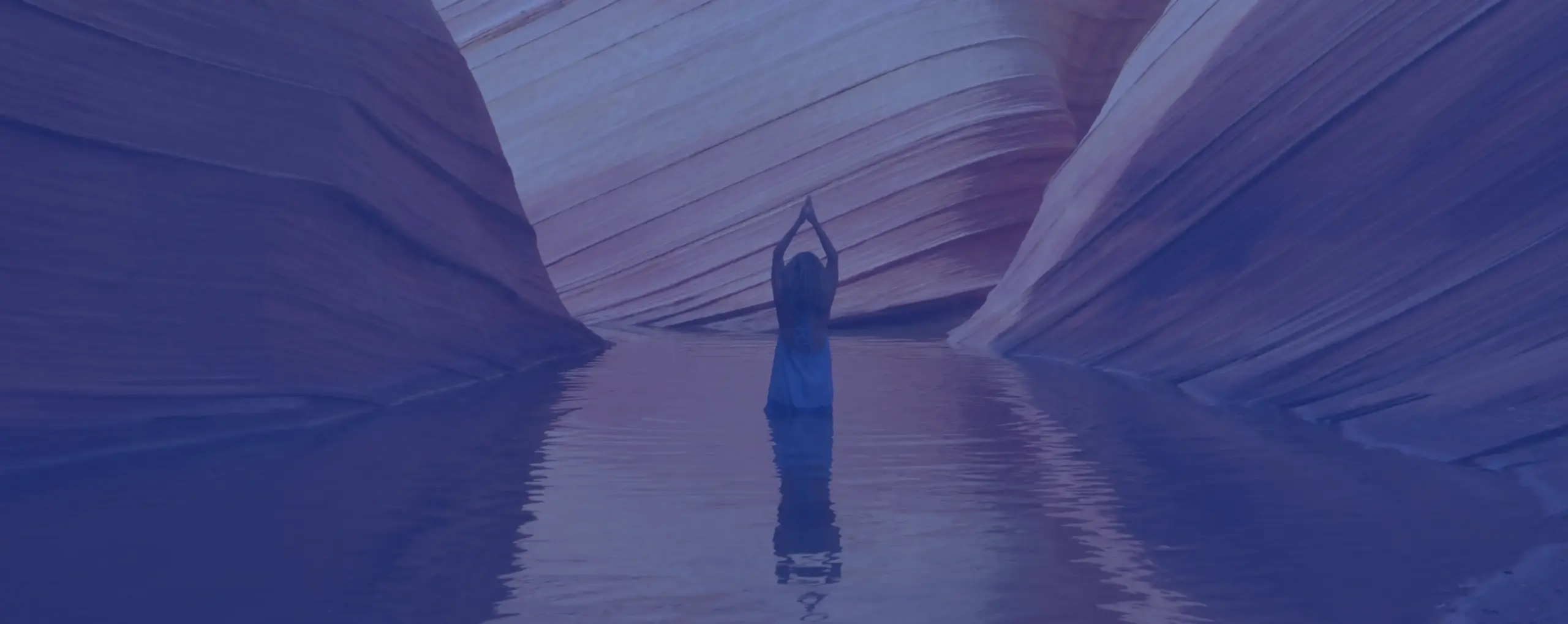 Person standing in shallow water between canyon walls with arms raised above head in a yoga pose.