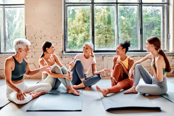 Group of diverse adults in athletic wear sits on yoga mats in a sunlit studio, chatting and laughing during a workout class.