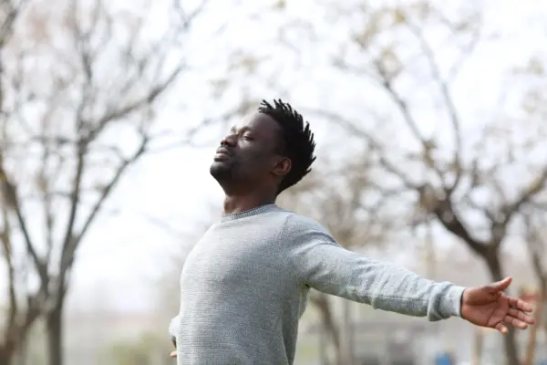 Man outdoors with arms outstretched and eyes closed, enjoying a breezy day in a park.