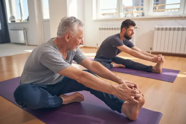 Two men wearing gray shirts on purple yoga mats, bending forward in a seated stretch in a sunlit studio.