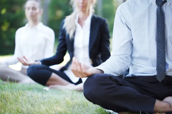 Three professionals meditating outdoors in a park; foreground man in a blue checkered shirt and tie sits cross‑legged with hands in lap.