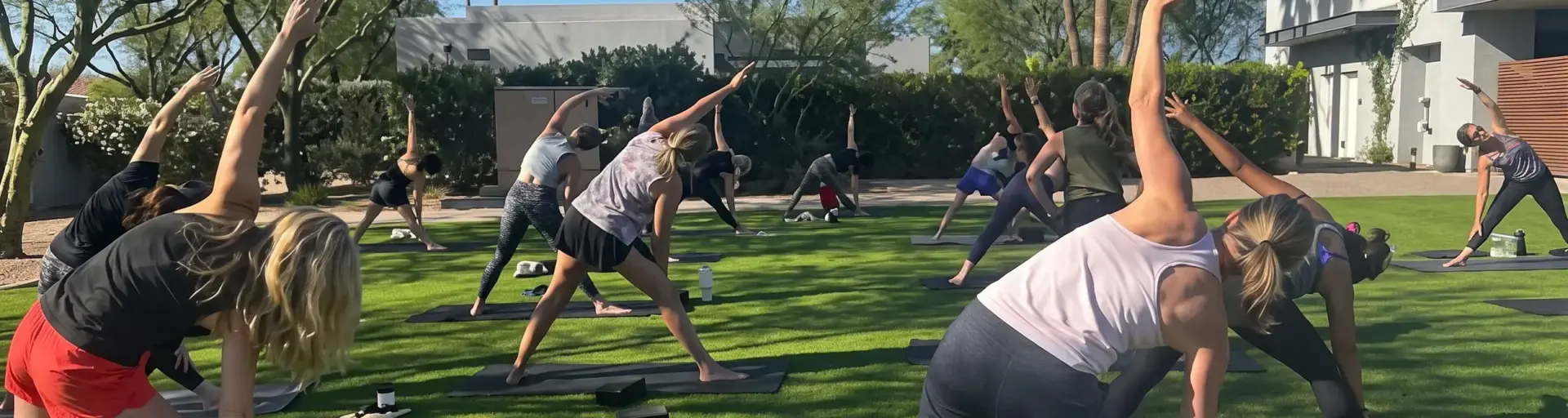 Group of people doing yoga on black mats in a sunny outdoor area with trees and modern buildings in the background.