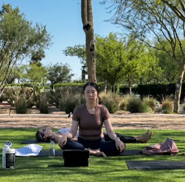 Woman meditating on a yoga mat outdoors with another person lying back on a mat nearby, trees and blue sky in background