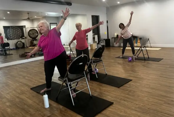 Three people doing arm-raised side stretches in a studio class, each on a yoga mat with mirrors on the wall behind them.